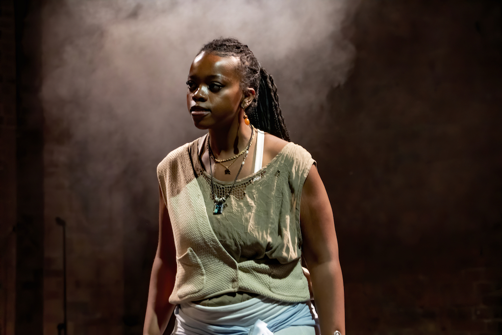 An actor stands alone on stage in mid-performance. Behind her mist from a smoke machine lingers in the air.
